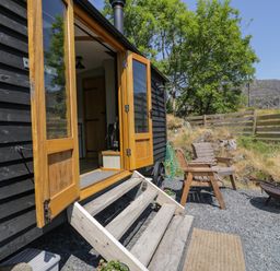 An outdoor seating area with wooden chairs and fire pit next to a black wooden building with open doors at Llethr - Shepherds Hut in Llanbedr