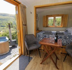 A small room with a bed a wooden table with wine glasses and a bottle two chairs and an open door leading outside to a hot tub at Llethr - Shepherds Hut in Llanbedr