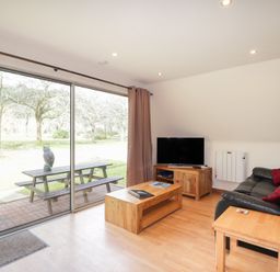 A living room with a black sofa red cushions wooden tables and a TV near large sliding glass doors at Owl Lodge South Laggan near Invergarry