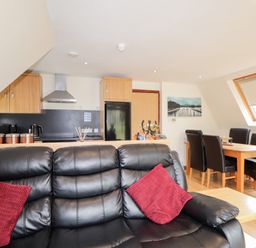 A living room with a black leather sofa and red cushions next to a wooden dining table with six dark chairs and a kitchen with wooden cabinets and black appliances at Owl Lodge in South Laggan near Invergarry