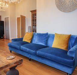 A blue sofa with yellow and blue cushions in a living room with a wooden table and kitchen area at 7 Four Seasons in Carbis Bay