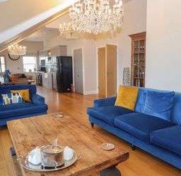 An open living room and kitchen area with blue sofas yellow cushions wooden coffee table and chandeliers at 7 Four Seasons in Carbis Bay