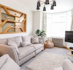 A living room with a grey sofa, a beige rug, a wall-mounted wooden shelf, a television on a stand, and a large window with patterned curtains at Dovestones in Keswick