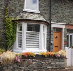 A stone house exterior with a bay window flowers and plants by the window and a wooden front door at Dovestones in Keswick