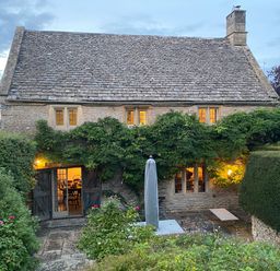 Stone cottage with lit windows and wooden door surrounded by greenery and garden furniture at The Small House Near Bourton-On-The-Water