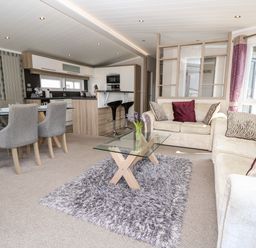 An open plan living room with beige sofas a glass coffee table on a rug dining table with chairs and a kitchen with stools at Crosswinds 67 in Bembridge