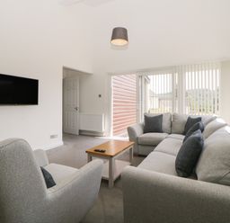 A living room with a gray armchair a gray sectional sofa with dark pillows a wooden coffee table a wall-mounted television a pendant light and large windows with vertical blinds at Harcombe House Bungalow 4 in Chudleigh