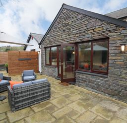 An outdoor patio area with stone paving a wicker sofa and chairs with cushions an umbrella and a stone wall house with windows and a door at Dant Y Llew in Bow Street Wales