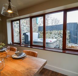 A dining area with a wooden table set with plates bowls and glasses next to a window showing a hot tub and stone wall outside at Dant Y Llew in Bow Street Wales