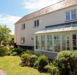 A garden with shrubs and trees beside a house with a glass conservatory at Home Farm Cottage in Fremington Devon