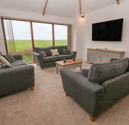 A living room with three gray sofas a coffee table a large window a wall-mounted television and a sideboard at Pol View in Gunwalloe near Helston