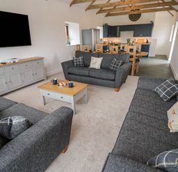 A living room with three gray sofas a wooden coffee table and a dining area with wooden chairs and blue cabinets at Pol View Gunwalloe near Helston