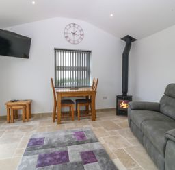 A living room with a grey sofa a small wooden dining table with two chairs a wall clock and a wood burning stove at Tyddyn Bach in Rhydwyn