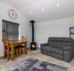 A small living room with a gray sofa a wooden table with two chairs a rug a black stove and a wall clock at Tyddyn Bach in Rhydwyn
