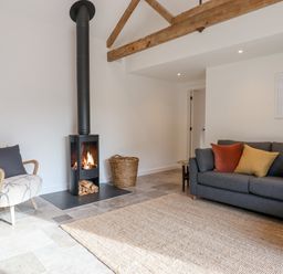 A living room with a wood burning stove a gray sofa with cushions an armchair and a woven basket at 1 Caddaford Barns in Buckfastleigh