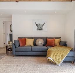 A living room with a grey sofa with orange and yellow cushions and a framed picture of a highland cow on the wall at 1 Caddaford Barns in Buckfastleigh