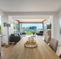 A dining table and chairs in an open living area with a sofa, armchair, and a view of houses and trees at Trewartha in Praa Sands