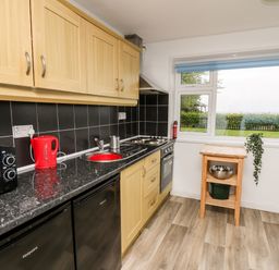 A kitchen with wooden cabinets black countertop a red kettle a microwave a stove and a small wooden table under a window at Cwtsh Cragen in New Quay