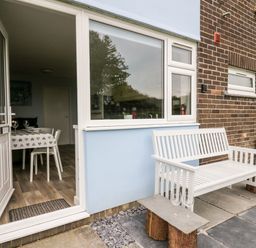 An outdoor patio area with a white bench and a small table holding two wine glasses next to a door opening to a dining area at Cwtsh Cragen in New Quay