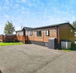 A wooden lodge with a small deck and steps with trees and water in the background at Gerrida Lodge South Lakeland Leisure Village near Tewitfield