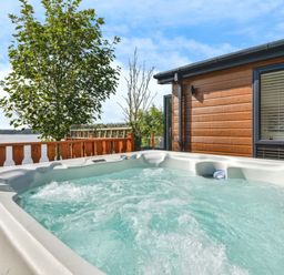 An outdoor hot tub on a wooden deck next to a cabin with a window and trees by a lake at Gerrida Lodge South Lakeland Leisure Village near Tewitfield