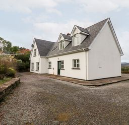 A white two-story house with a sloped roof and windows on a gravel driveway with surrounding greenery at Seascape Lodge in Kenmare County Kerry