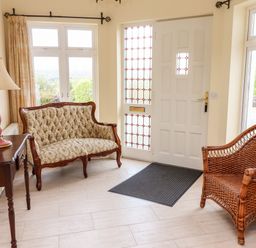 An entryway with a floral upholstered wooden settee a wicker chair a wooden table with a lamp and a white door with stained glass panels at Seascape Lodge in Kenmare County Kerry