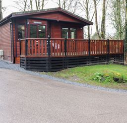 An exterior view of a wooden cabin with a porch surrounded by trees at Alfresco Lodge in Skiptory Howe 37B White Cross Bay near Windermere