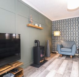 A living room with a television on a wooden stand a black stove a blue armchair and patterned wallpaper at Alfresco Lodge Skiptory Howe 37B White Cross Bay near Windermere