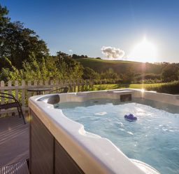 An outdoor hot tub on a deck with two chairs and a table overlooking a grassy hill at Apple Barn in South Molton