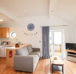 A living room and kitchen area with a grey sofa wooden cabinets a clock and a TV at Beside the Sea in Eype