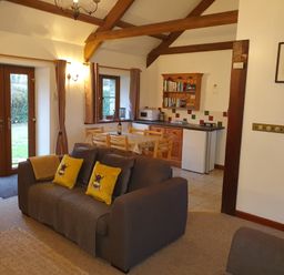 A living area with a gray sofa with yellow pillows a dining table with chairs a small kitchen with wooden cabinets a bookshelf a microwave and double glass doors at Ty Barcud in Llanfair Clydogau