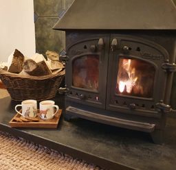 A wood burning stove with fire inside a basket of firewood and two mugs with biscuits on a tray at Ty Barcud in Llanfair Clydogau
