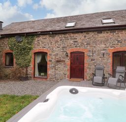 An outdoor area with a white jacuzzi a wooden deck with two gray reclining chairs and a stone building with red brick window frames at The Hawthorns in North Molton