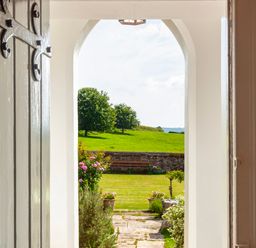 A view through an open door to a garden with flowers a stone path a bench and green fields at Crablake Farmhouse in Exminster