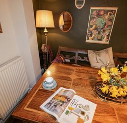A wooden dining table with a magazine a cup and saucer and a vase with yellow flowers against a dark wall with mirrors and framed art at The Nook in Whitby