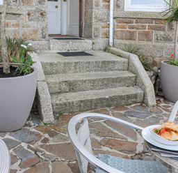 An outdoor stone patio with a table set with plates and croissants and two large plant pots on either side of stone steps leading to a door at Whistler in St Ives