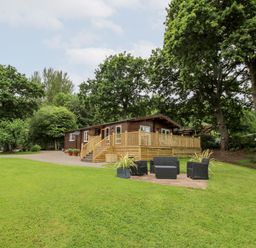 A cabin with a wooden deck and outdoor seating area on a grassy lawn surrounded by trees at The Log Cabin in Newnham Bridge near Tenbury Wells