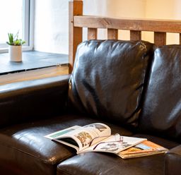 A black leather sofa with magazines and reading glasses on the seat near a window with a small potted plant at Stable Cottage in Port Isaac