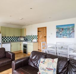 An open living space with a kitchen area including green cabinets and checkerboard backsplash a dining table with white chairs and dark brown leather sofas in the foreground at Gull Lodge in Port Isaac