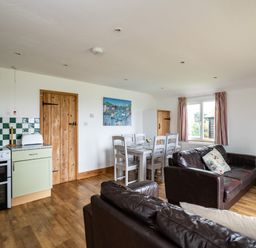 A living room with brown leather sofas a dining table with chairs and a kitchen area with a stove and wooden doors at Gull Lodge in Port Isaac