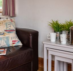 A leather couch with a nautical themed pillow and glasses on a book next to nested white tables with mugs a metal coffee pot and plants at Gull Lodge in Port Isaac