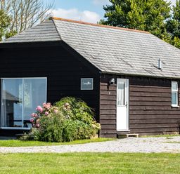 A black wooden cottage with a gray tiled roof flowers in front and a picnic bench on the side at Campion Lodge in Port Isaac