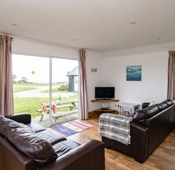 A living room with two dark brown sofas a small television on a corner shelf a window and sliding glass door with curtains at Campion Lodge in Port Isaac