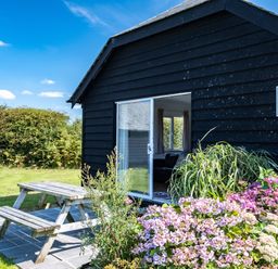 A black wooden building with a sliding door opening to a room with a picnic table outside and flowers in front at Harbour Lodge in Port Isaac