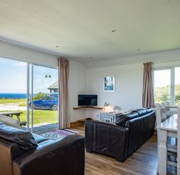 A living room with two dark sofas a corner TV table a white dining table with chairs and a large window with a sea view at Harbour Lodge in Port Isaac