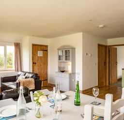 A living room and dining area with a white table set with plates and bottles a gray cabinet two wooden doors and a kitchen area visible at Puffin Lodge in Port Isaac