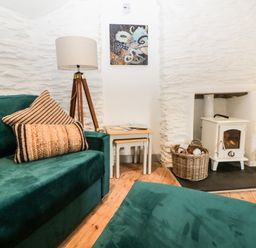 A room with a green sofa a floor lamp a wood stove with tools and basket tables and artwork on a white textured wall at Spindrift in Polperro