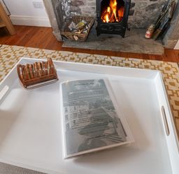 A white tray on a patterned rug with a booklet and a wooden napkin holder in front of a fireplace with a burning fire at Cross View Cottage in Alston