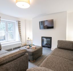 A living room with grey sofas a window with curtains a wall-mounted TV and an electric fireplace at Beach Road House in Hele Bay Ilfracombe
